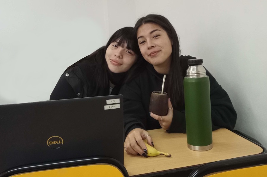 En un aula de la Facultad de Informáticas dos alumnas del taller posan para la foto. Una de ellas tiene un mate y una banana en la mano. Hay un termo de color verde en el escritorio y una notebook.